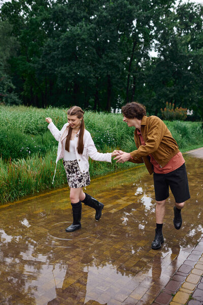 A cheerful young couple walks hand in hand along a puddle filled path in a lush park.