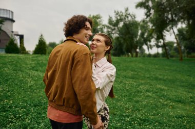 Genç çift, yemyeşil bir parkta sakin bir yürüyüş yaparken sevgi dolu bakışlarını paylaşıyor..