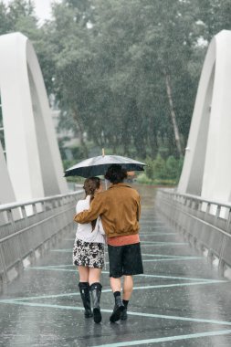 Genç çift yağmurda birlikte dolaşır, yemyeşil bir manzaranın ortasında şemsiye altında sıcaklığını paylaşır..