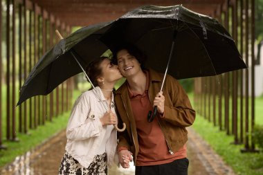 Genç aşıklar, hafif yağmur altında parkta yürürken samimi bir anı paylaşırlar..