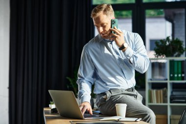 Modern ofis ortamında dizüstü bilgisayar üzerinde çalışırken genç bir profesyonel telefonla konuşuyor..