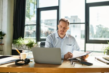 Odaklanmış genç bir işadamı, güneşli bir ofiste modern bir masada otururken belgeleri inceliyor..