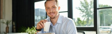 Genç bir işadamı modern bir ofiste oturur, kahveyi yudumlarken gülümser ve kendine güvenir..