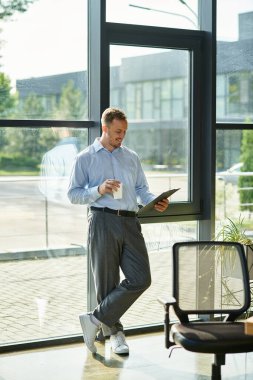 Geniş bir ofis penceresinin yanındaki genç profesyonel, kahvesini yudumluyor ve notlarını gözden geçiriyor..