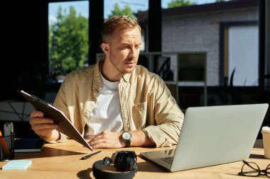 Şık bir ofisteki genç adam elinde derin düşüncelerle elinde bir belge tutarken dizüstü bilgisayarına odaklanıyor..