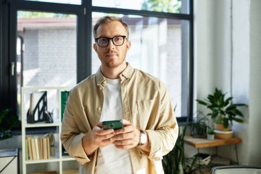 Genç bir profesyonel kendine güvenerek şık bir ofiste duruyor, akıllı telefonuna dalmış..