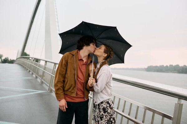Молодая пара нежно целуется, идя по мосту под легким дождем, обнимая любовь.