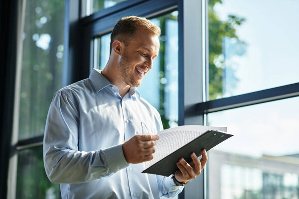 A handsome young professional smiles while checking important paperwork in a sunlit office.
