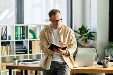 Yakışıklı genç profesyonel iyi aydınlatılmış bir ofiste dururken bir deftere notlar yazar..