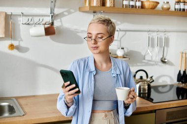 Kısa saçlı genç bir kadın evde rahatlıyor, kahvesini yudumluyor ve telefonunu kullanıyor, sakince yayılıyor..
