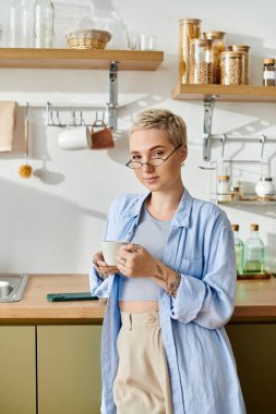 Kısa saçlı güzel bir kadın evde dinleniyor, parlak bir mutfakta kahvenin tadını çıkarıyor..