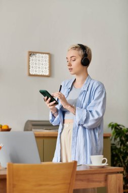 Kısa saçlı genç bir kadın evde kulaklık takarken telefonunu kullanıyor..