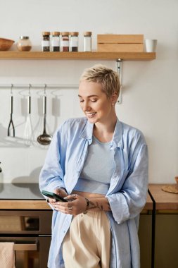 Kısa saçlı genç bir kadın ev mutfağında akıllı telefonunu kullanırken gülümsüyor..