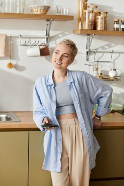 Modern mutfağında rahat ve mutlu görünürken kısa saçlı bir kadın gülümsüyor.