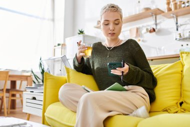 Güzel genç bir kadın parlak sarı bir kanepede dinleniyor, not yazarken çayını yudumluyor..