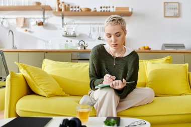 Genç bir kadın modern bir evde parlak sarı bir kanepede otururken düşünceli bir şekilde plan yapar..