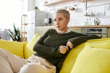 Kısa saçlı genç bir kadın güneşli sarı bir kanepede rahatça oturur, düşüncelere dalmış..