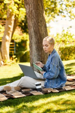 Laptopunda kısa saçlı, ağaçlarla çevrili genç bir kadın..