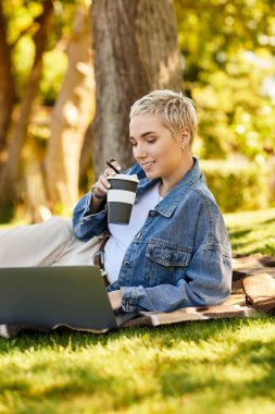 Genç bir kadın güneşli bir parkta battaniyeye uzanıp laptopuna odaklanıyor ve kahvesini yudumluyor..