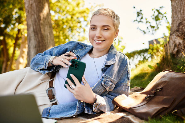 A young woman with short hair relaxes outside, smiling while using her phone on a sunny day.