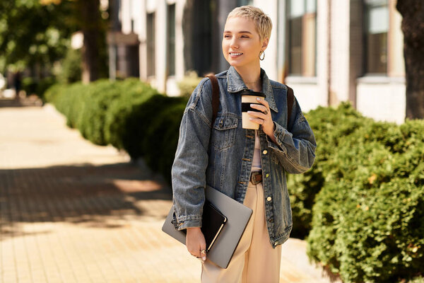 A young woman with short hair walks outdoors, holding a coffee cup and a laptop bag while smiling.