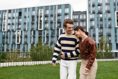 İki genç aşık birbirine yakın yürüyor, çağdaş binalarla dolu bir şehir parkında kahkahalarda kayboldular..