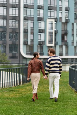 Sevgi dolu çift, çağdaş binaların yakınındaki canlı yeşil bir alanda el ele yürüyor..