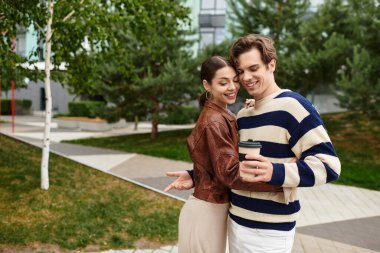 Genç bir çift, dışarıda kahve fincanları tutarken sıcak bir kucaklaşmayı ve neşeli bir gülümsemeyi paylaşıyor..