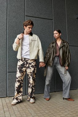 Romantik bir çift, modern bir arka plana karşı eşsiz moda anlayışlarını sergilerken el ele tutuşur..