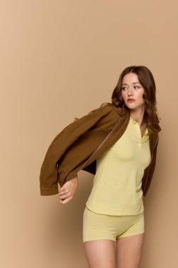 Güzel genç bir kadın, bej bir arka fona karşı kahverengi ceketli şık bir sarı elbiseyle poz veriyor..