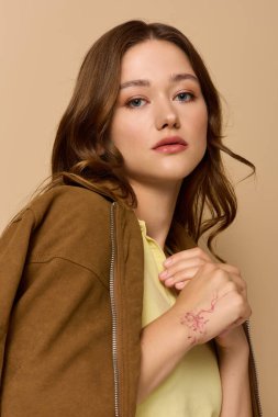 Genç bir kadın şık sarı bir elbise ve bej bir stüdyoda kahverengi bir ceket içinde zarif poz veriyor..