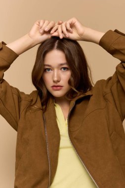 Genç bir kadın, bej bir arka fona karşı kahverengi ceketli şık sarı bir elbise içinde poz veriyor..