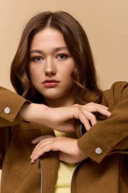 Sarı elbiseli ve kahverengi ceketli kendine güvenen genç bir kadın bej bir arkaplanda poz veriyor..