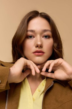 Genç bir kadın bej bir stüdyoda kahverengi ceketli şık sarı bir kıyafet sergiliyor..