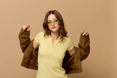 Güzel bir kadın, bej bir stüdyo ortamında kahverengi ceketli, sarı bir elbiseyle rahatça poz verir..