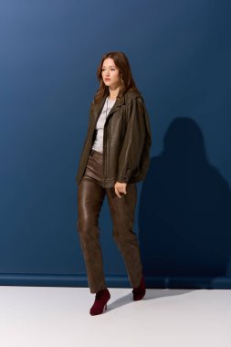 Genç bir kadın, şık mavi bir stüdyoda sonbahar atmosferini yakalayan kahverengi deri bir elbise mankenliği yapıyor..