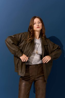 Genç ve güzel bir kadın, kahverengi deri bir elbiseyle canlı mavi bir arkaplana karşı güvenle poz veriyor..