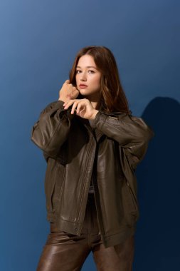 Genç bir kadın kahverengi deri bir elbiseyle mavi bir stüdyo arkaplanına karşı güvenle poz veriyor..