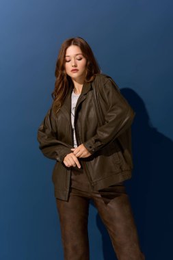 Genç ve güzel bir kadın canlı mavi bir stüdyo ortamında şık bir kahverengi deri elbise sergiliyor..