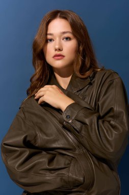 Modaya uygun genç bir kadın, mavi bir stüdyo ortamında kahverengi deri bir elbiseyle rahatça poz veriyor..