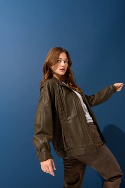 Güzel genç bir kadın, sonbahar modasını mavi bir stüdyoda kahverengi deri elbisesiyle sergiliyor..