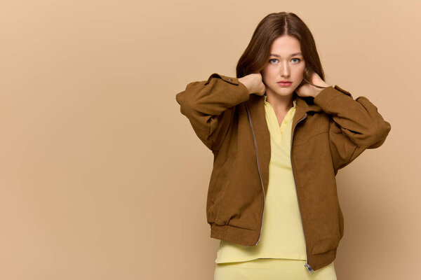 Молодая женщина уверенно позирует в стильном желтом наряде с коричневой курткой.