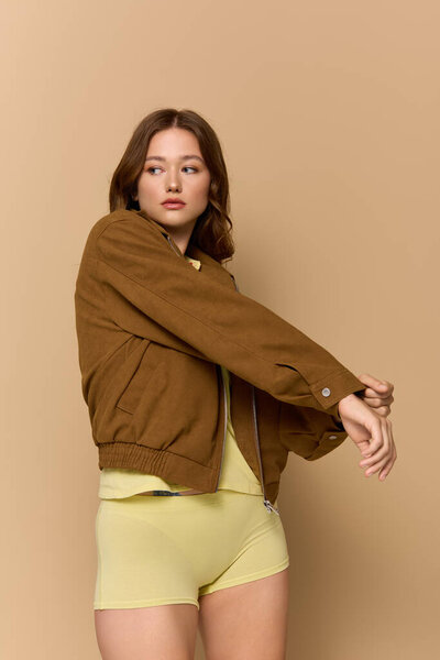 Confident young woman showcases a chic yellow outfit with a brown jacket in a stylish studio.