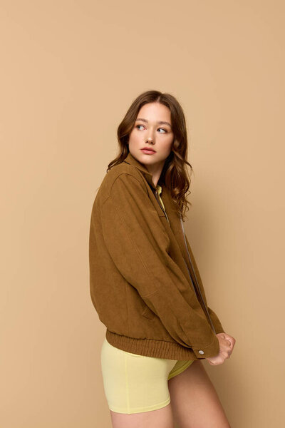 Pretty young woman showcases a trendy yellow outfit with a brown jacket in a studio setting.