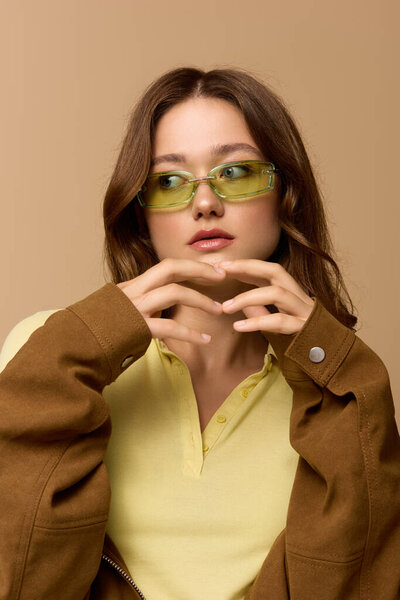 Pretty young woman in a yellow outfit and brown jacket poses confidently in a beige studio.