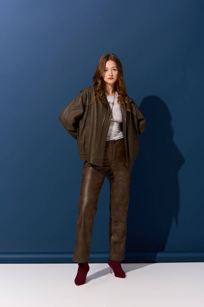Красивая молодая женщина уверенно позирует в коричневой кожаной одежде на синем фоне.