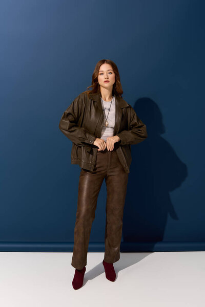 Красивая молодая женщина уверенно позирует в коричневой кожаной одежде на синем фоне.