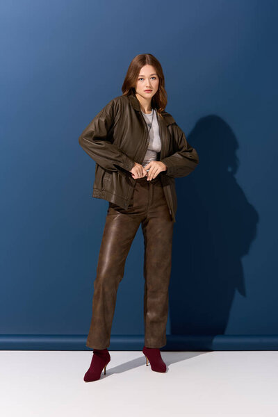 A young woman shows off her fashionable brown leather outfit against a vibrant blue backdrop.