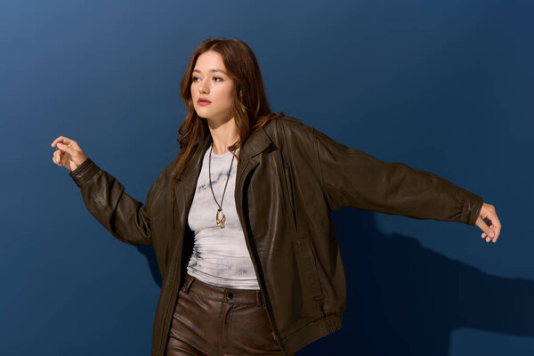 Pretty young woman poses confidently in a brown leather outfit against a blue background.