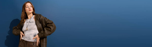 Молодая женщина уверенно позирует в коричневой кожаной одежде в синей студии, обнимая осень, баннер
